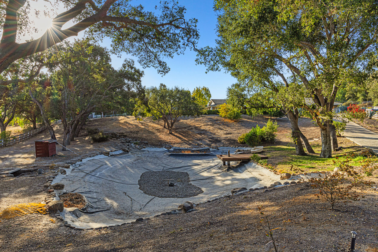 Landscaped yard with mature trees, a dry lined pond, and a small wooden footbridge.