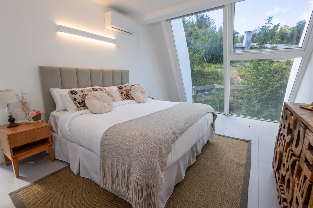 Bedroom with a white bed and large angled windows overlooking greenery.