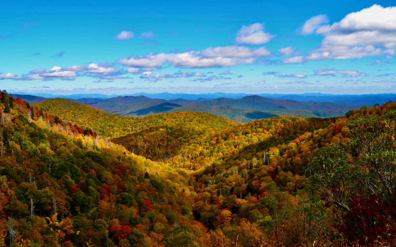 Layered mountain ridges and autumn-colored forest beneath a blue, cloud-dotted sky.