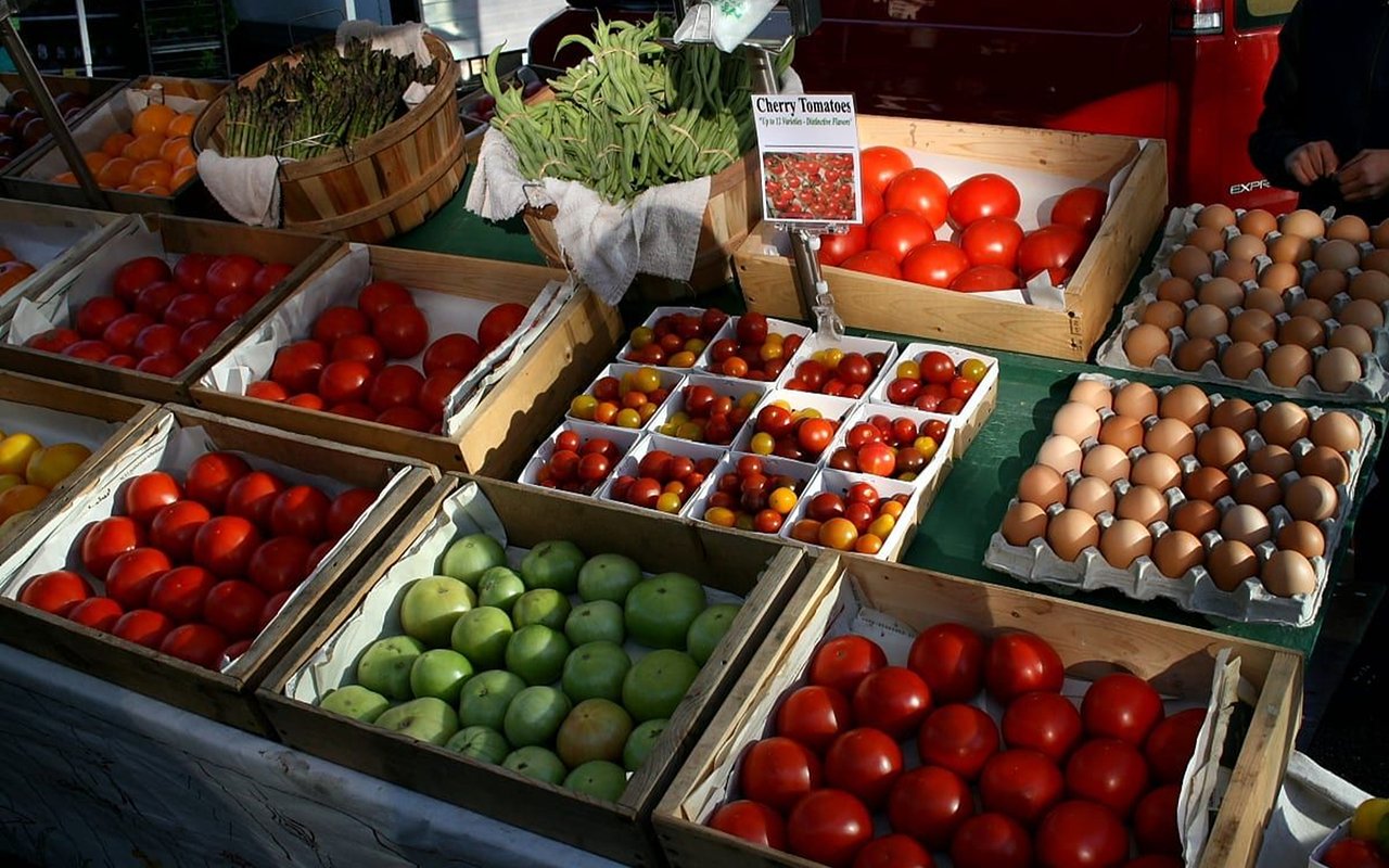 Market display of tomatoes, green beans, leafy greens, and eggs in crates and trays.