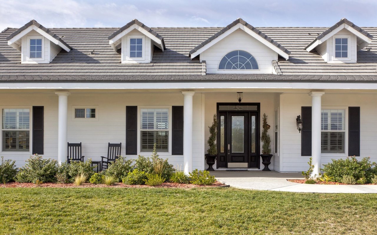 White house with gray tiled roof, multiple dormers, and a covered porch with a dark double front door.