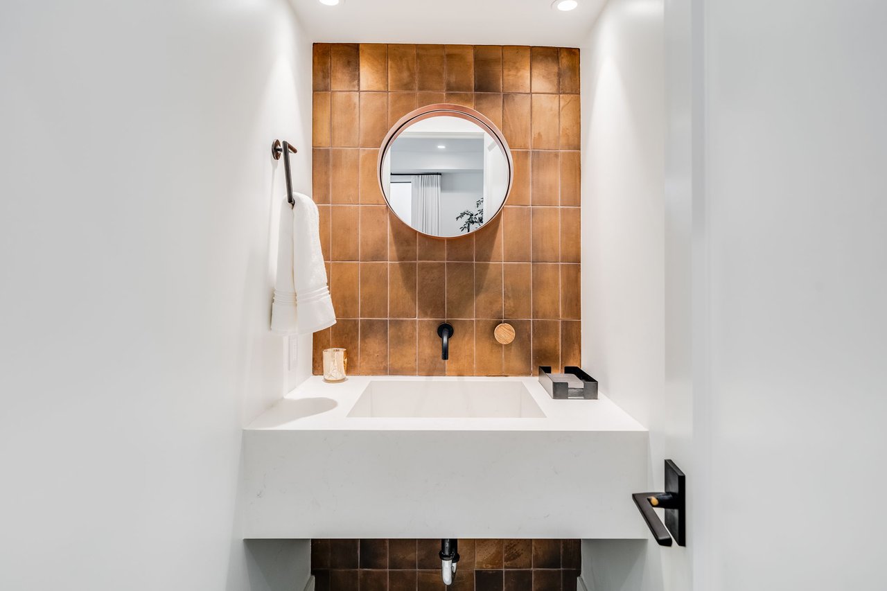 Bathroom vanity with a round mirror, copper-brown tiled backsplash, and matte black fixtures.