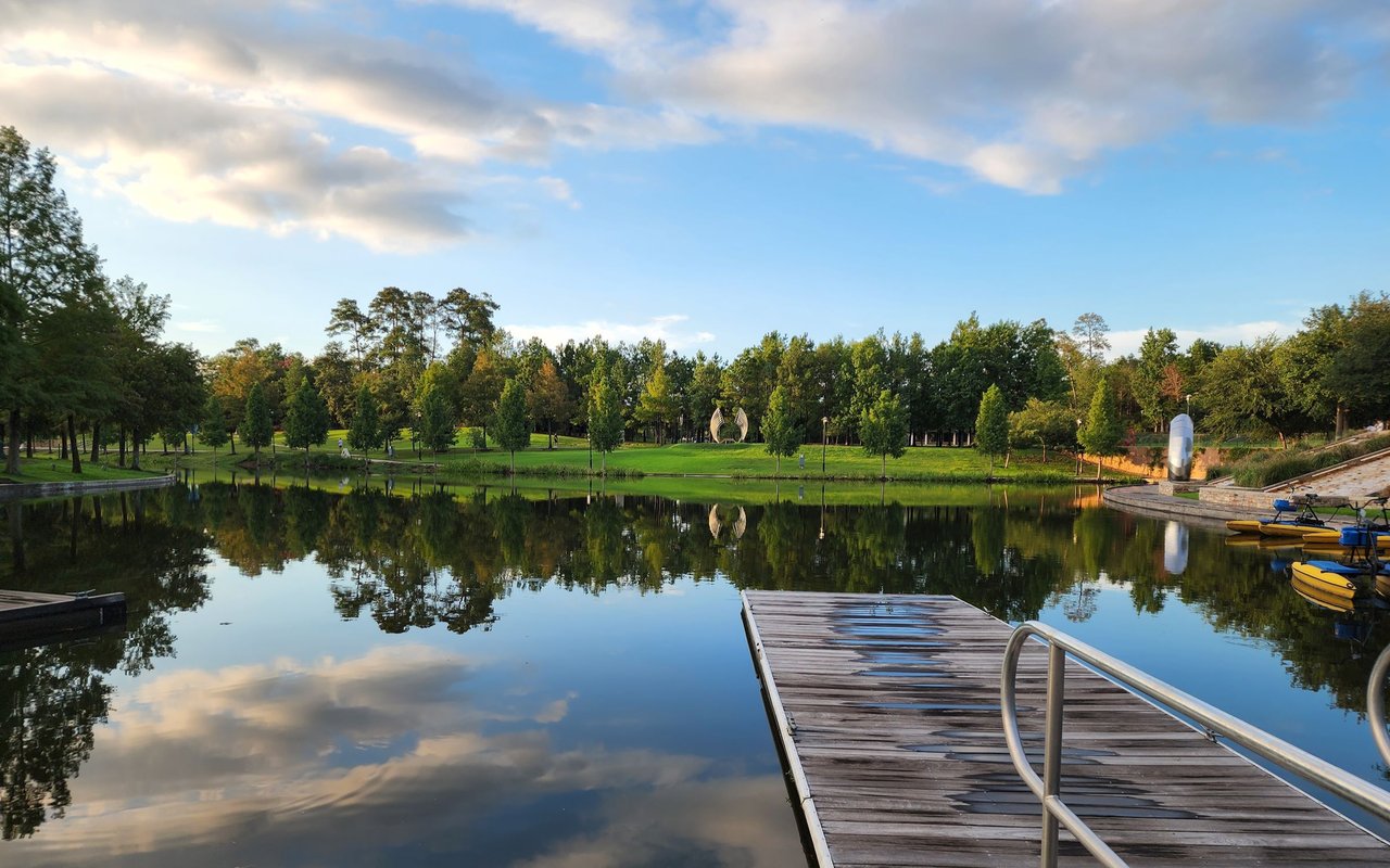Wooden dock extending into a calm, tree-lined lake with kayaks along the shore.