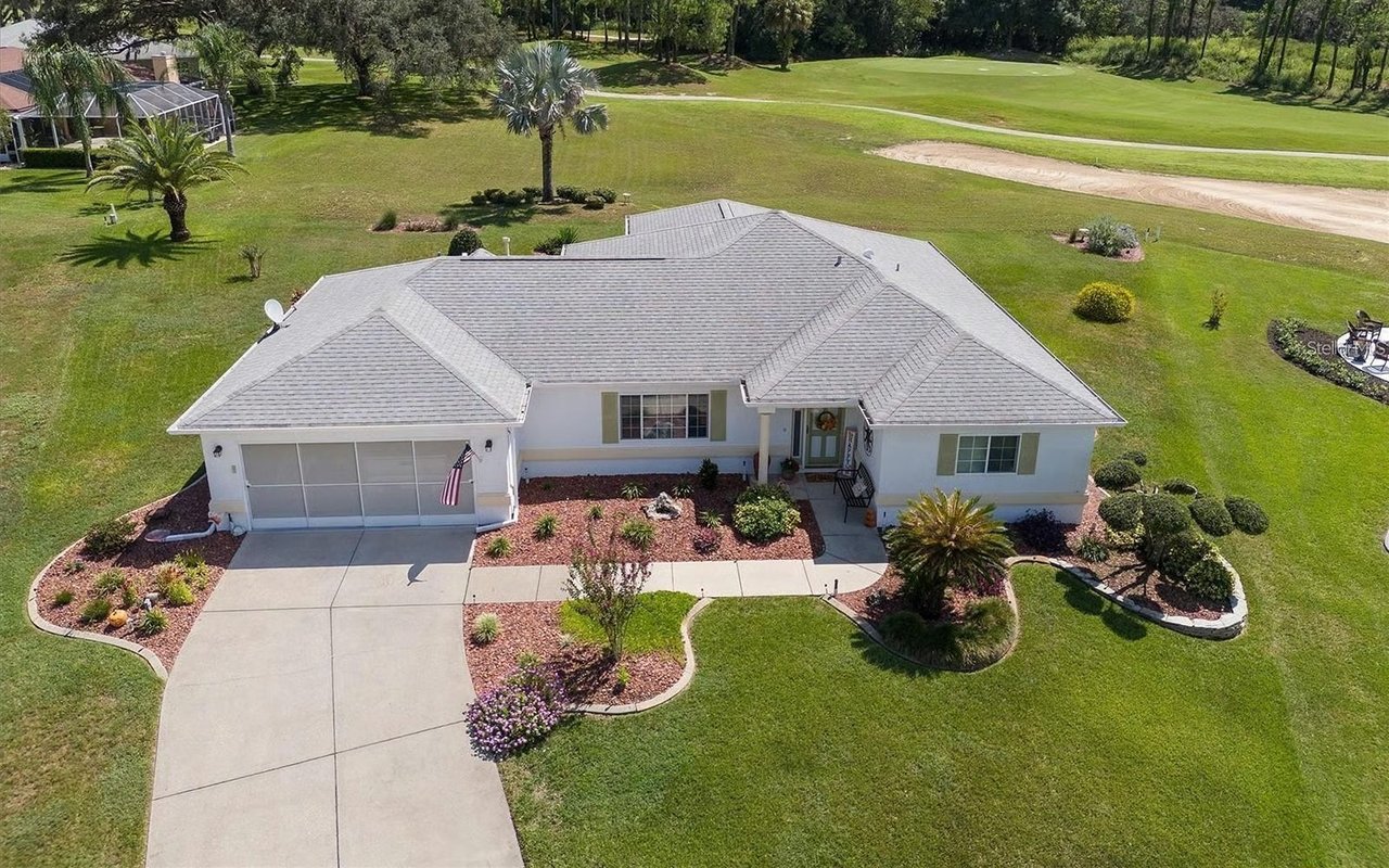 Aerial view of a white single-story home beside a golf course, with a broad driveway and landscaped front yard.
