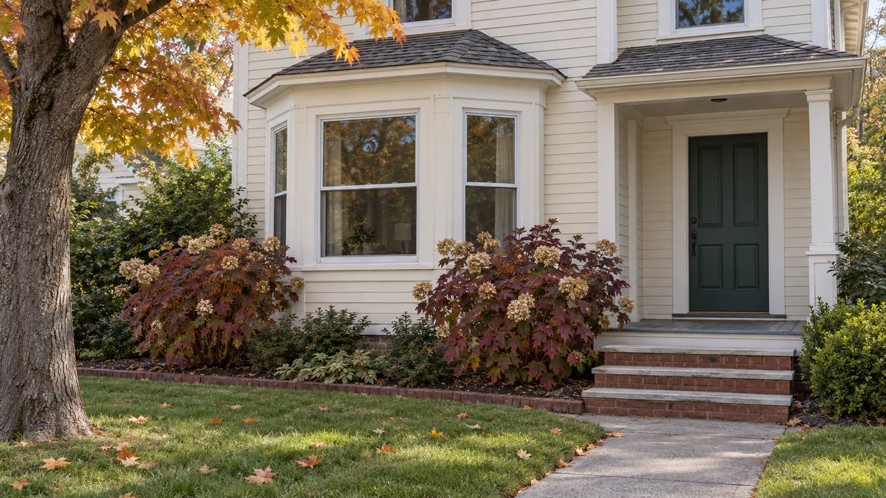 Ivory clapboard home with a projecting window bay, green front door, brick entry steps, and an autumn garden.