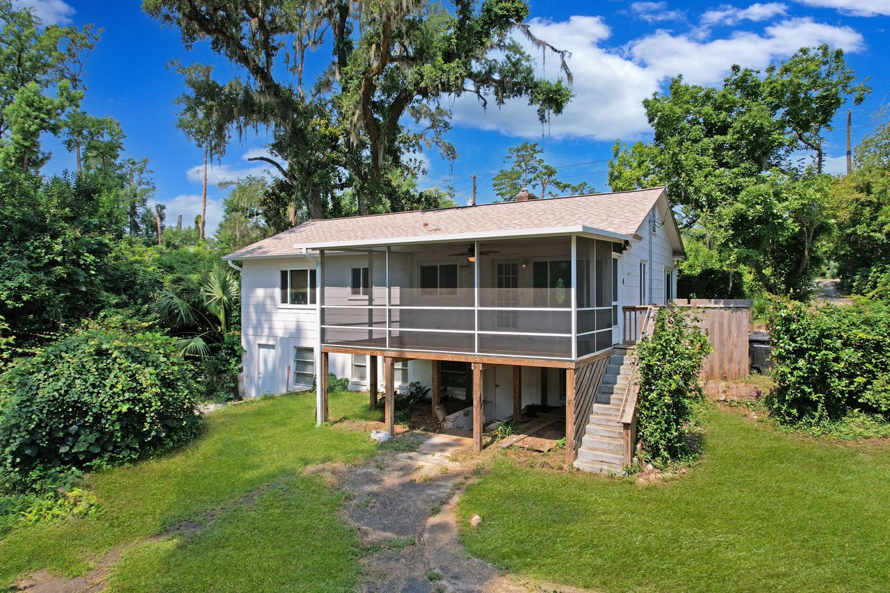 Elevated white house with a screened porch, surrounded by lush green trees and grass. The setting is peaceful, conveying a rustic, serene vibe.