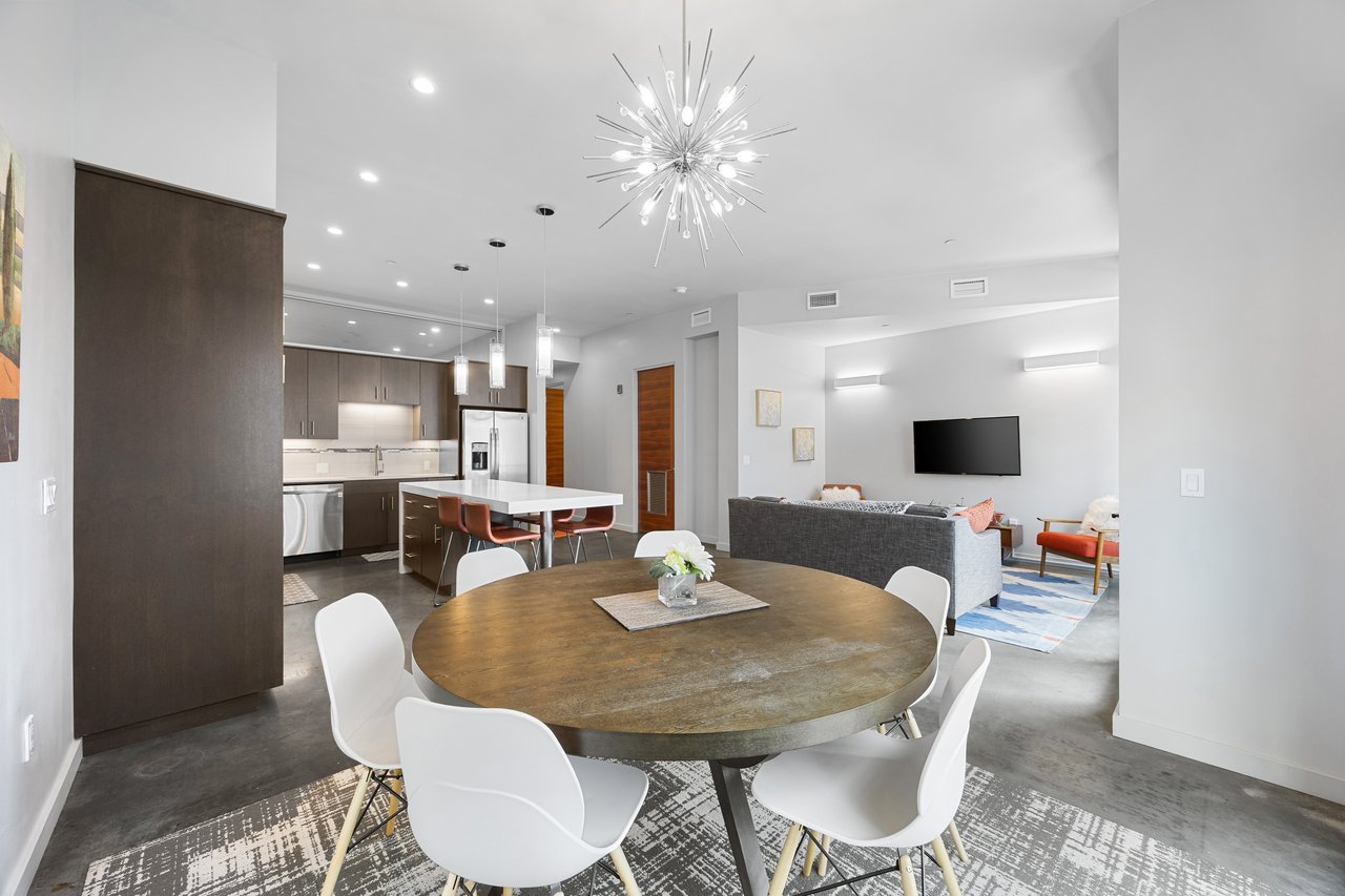 Open-plan dining area with a round wooden table, modern kitchen, and gray sofa beneath a starburst chandelier.
