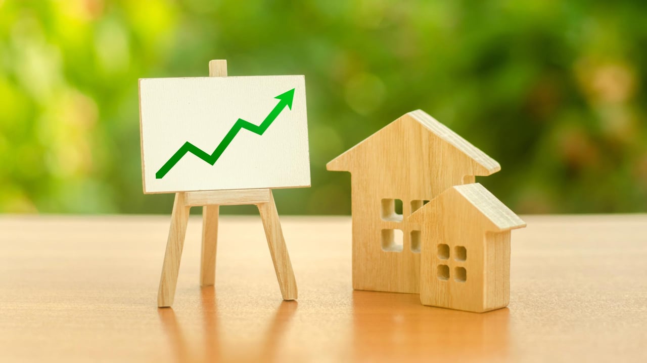 A descriptive close up of small wooden house models sitting next to a miniature easel displaying a sharp green upward trending arrow graph, illustrating the data driven real estate pricing strategy required when navigating a shifting property landscape and deciding how to price your home to secure maximum competitive engagement when selling a home GTA families value.