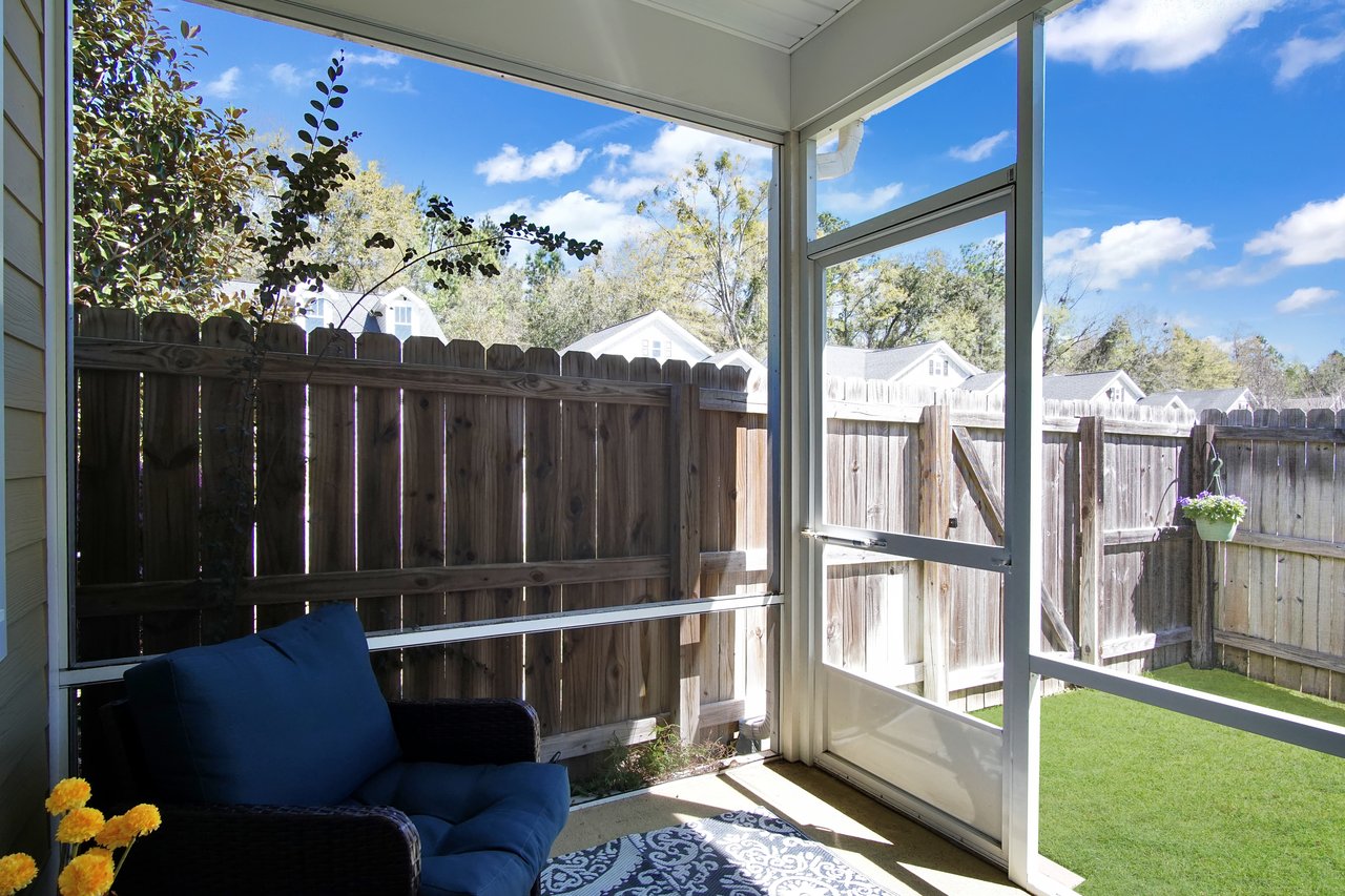 Sunny patio with wicker chair and blue cushions, overlooking a wooden fence and lush grass. Potted flowers hang near a clear blue sky. Inviting and serene.