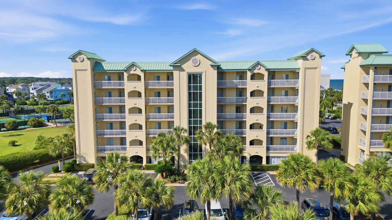 Multistory beige condominium with green roofs, stacked balconies, and palm trees in front.