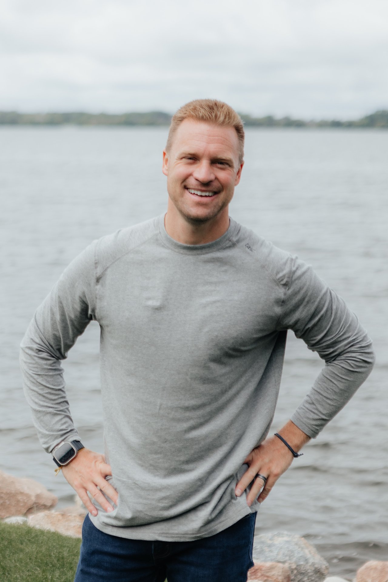 Smiling man in a gray shirt standing beside a lake with his hands on his hips.