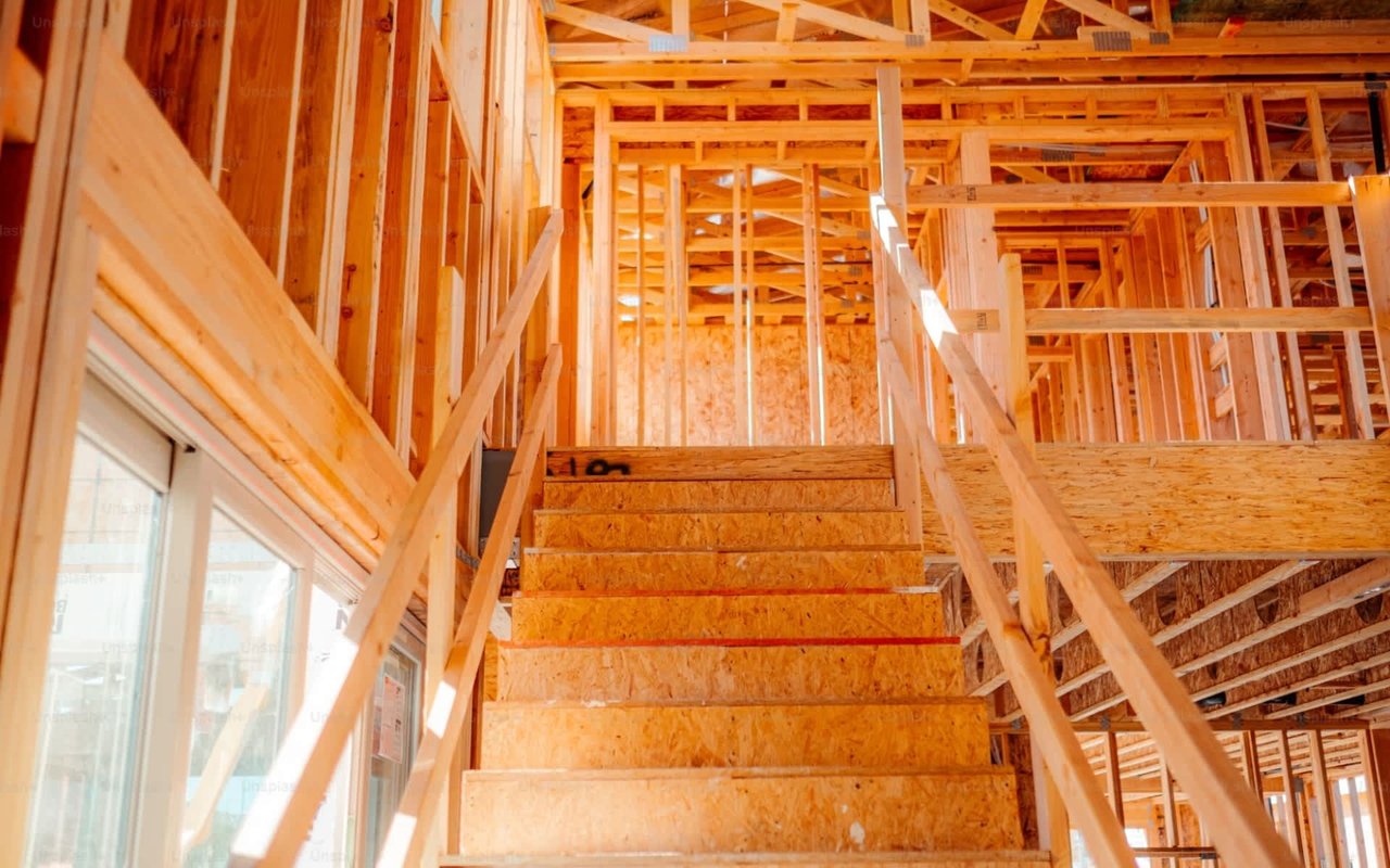 Interior framing of a house with exposed wooden studs, ceiling rafters, temporary handrails, and a central plywood staircase under bright light.