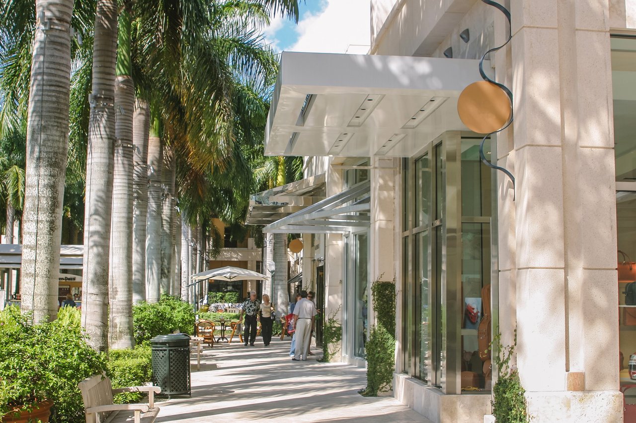 Palm-lined shopping promenade with glass-fronted shops, benches, and pedestrians.