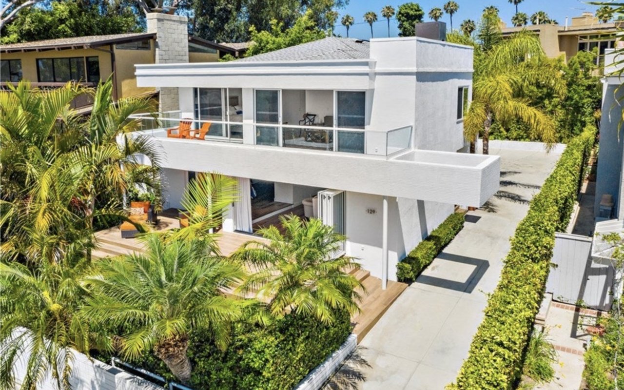 Modern white two-story home with a glass-railed balcony, palm-filled patio, and hedge-lined driveway.