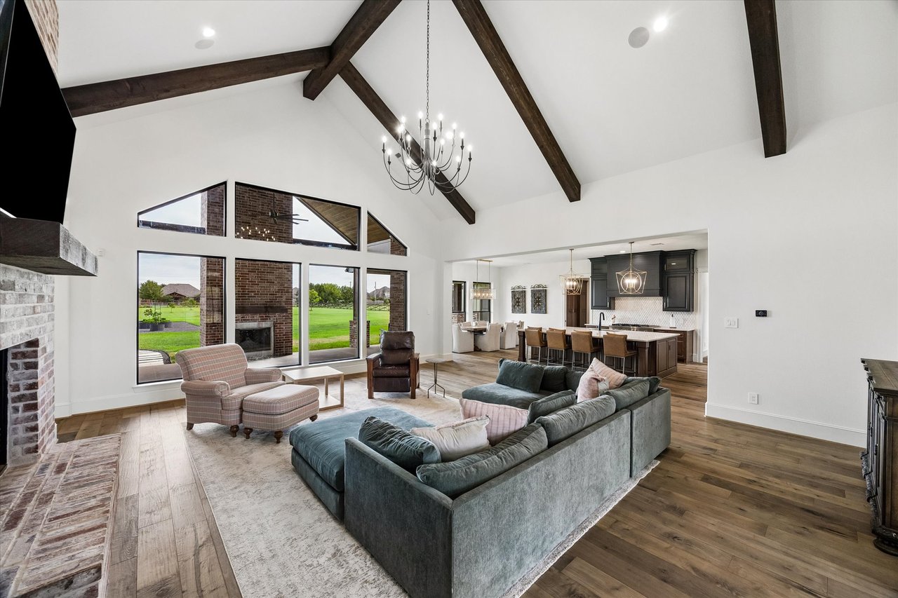 Living room with vaulted beamed ceiling, brick fireplace, and tall windows overlooking a lawn.
