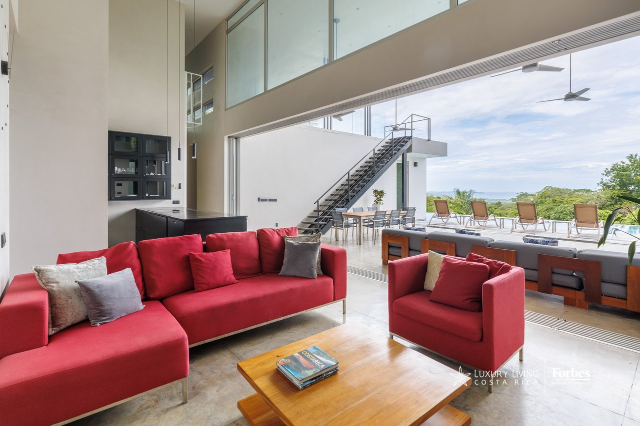 Double-height living room with red sofas, open glass doors, and a terrace overlooking greenery and water.