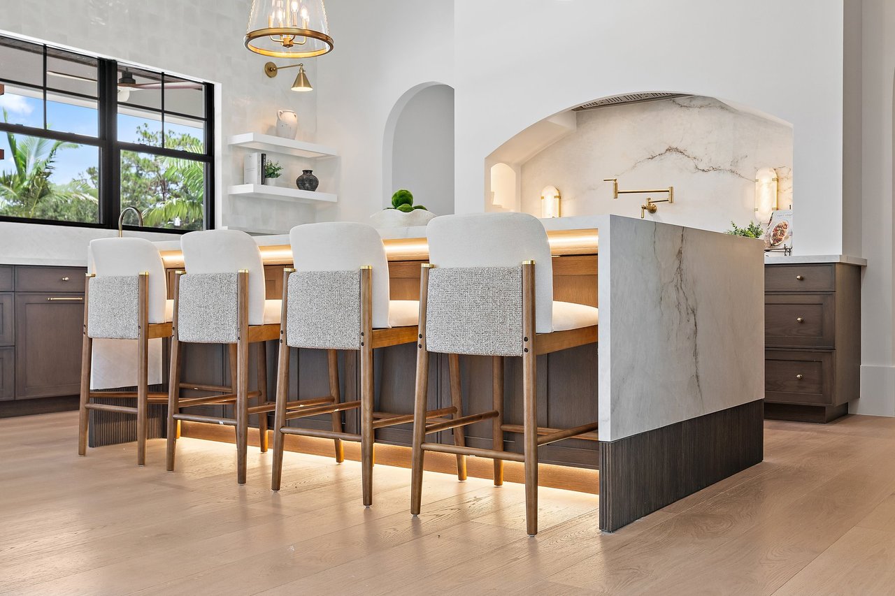 Modern kitchen with a marble island, four upholstered bar stools, and black-framed windows overlooking greenery.