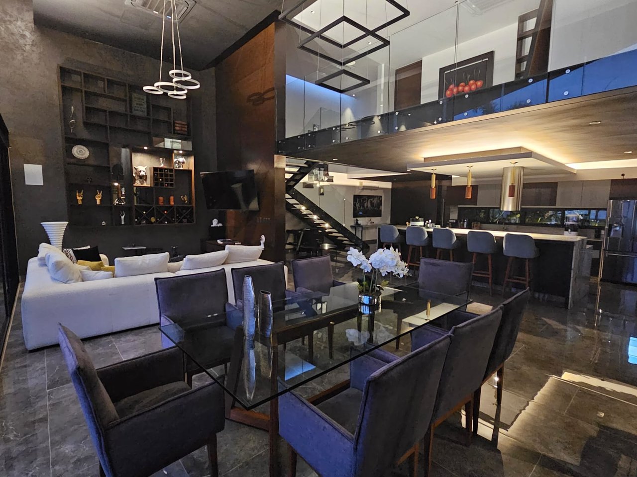 Double-height living and dining area with glass-railed mezzanine and illuminated staircase.
