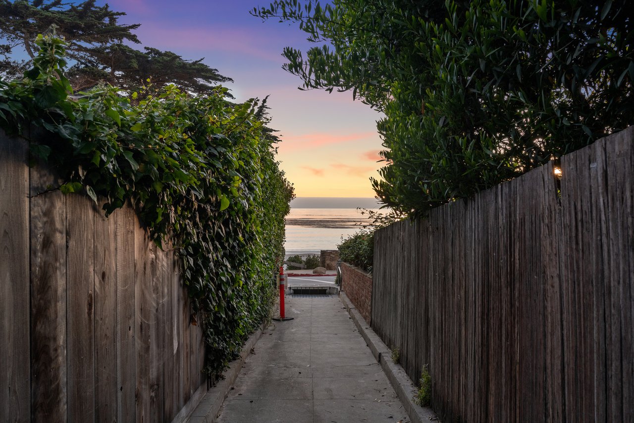 A quiet pathway lined with wooden fences and lush greenery leading down to Carmel Beach at twilight, with a colorful sunset sky over the ocean in Carmel-by-the-Sea, California