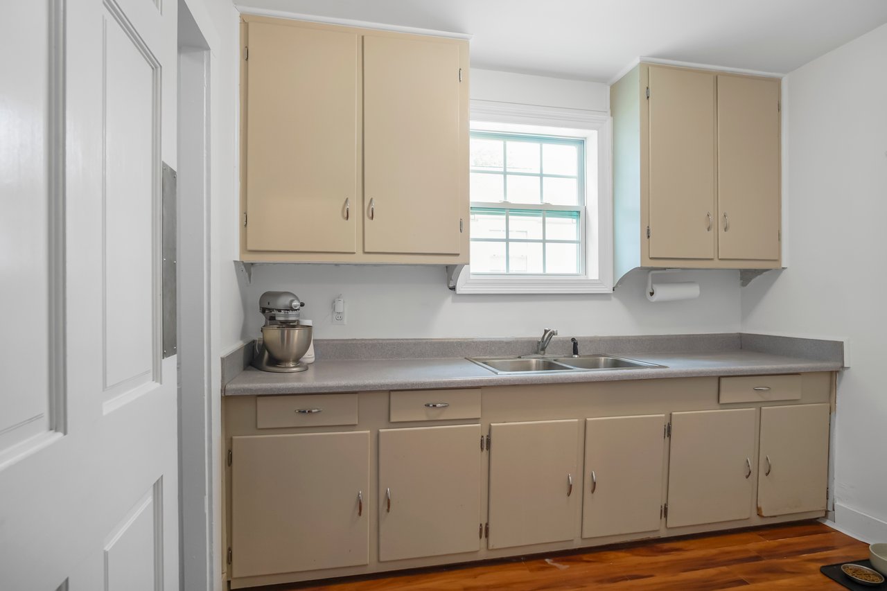 A small, simple kitchen with beige cabinets, a gray countertop, and a stainless steel mixer. A window above the sink lets in soft natural light.