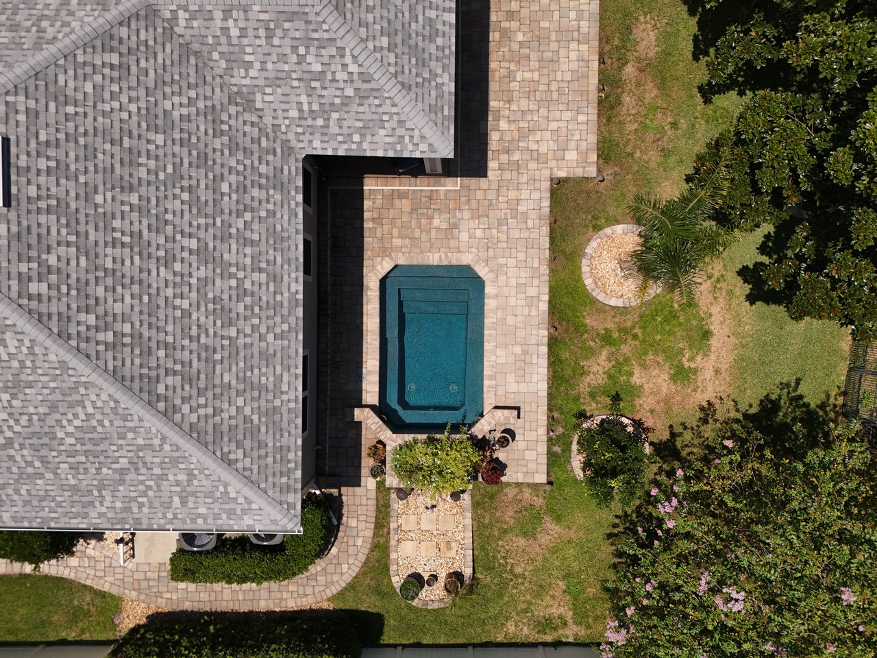 Aerial view of a backyard pool surrounded by a paved patio, lawn, and mature landscaping.