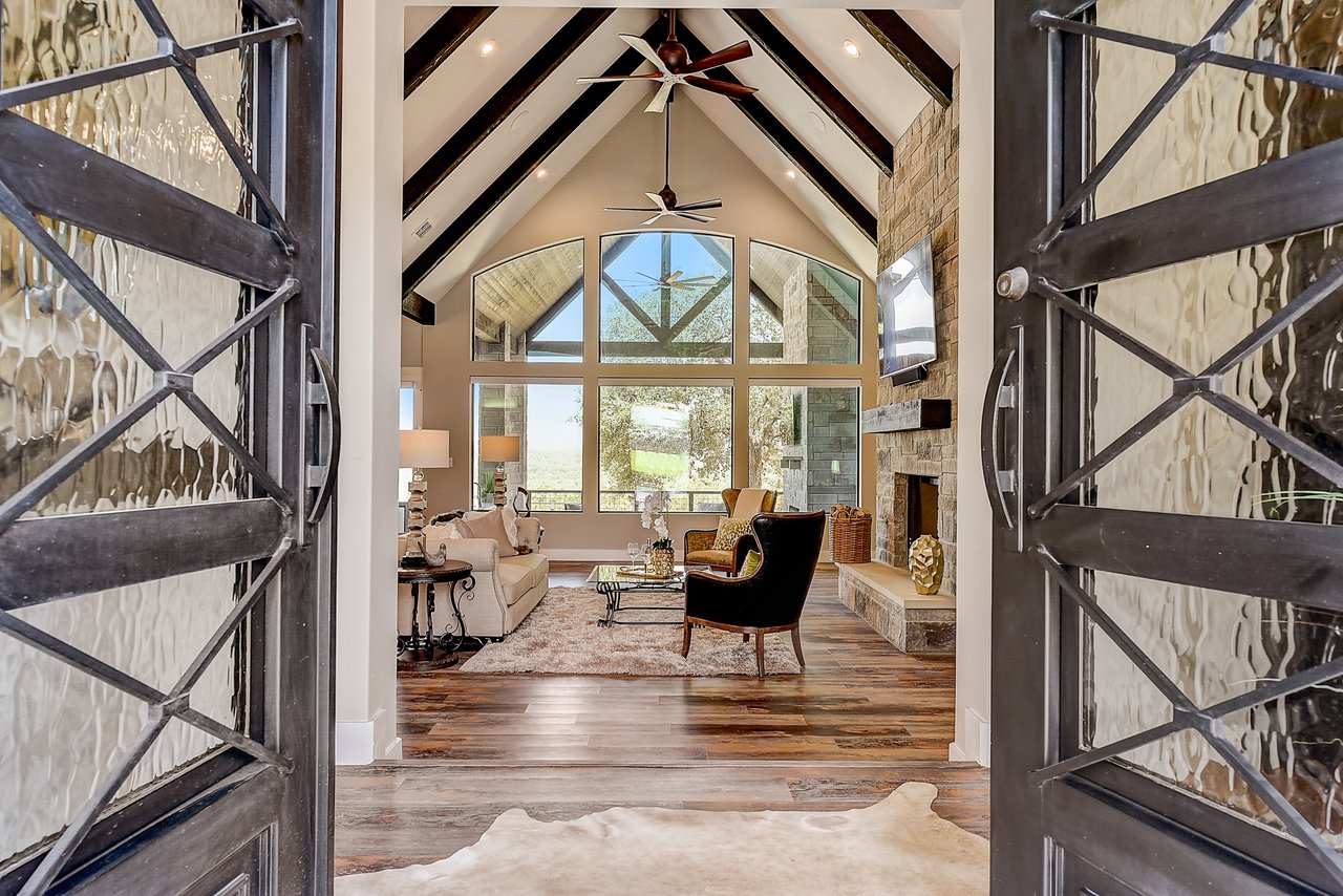 Iron and glass entry doors open to a vaulted great room with exposed beams, a stone fireplace, and Hill Country views in New Braunfels.