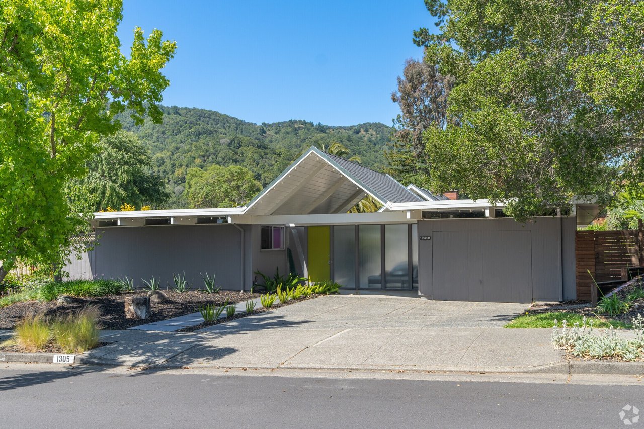 Modern gray home with a sharply peaked roof, yellow-green door, and wooded hills behind it.