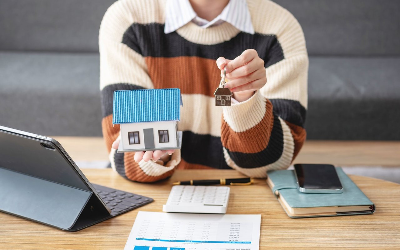Person holding a small house model and house-shaped key charm at a desk.