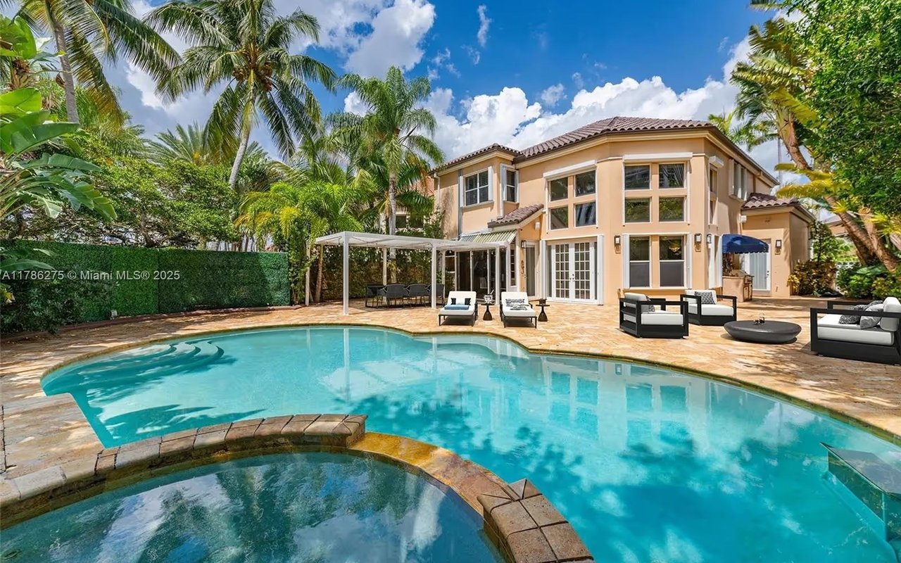 Two-story peach home behind a turquoise pool and raised spa, with palm trees and a furnished stone patio.