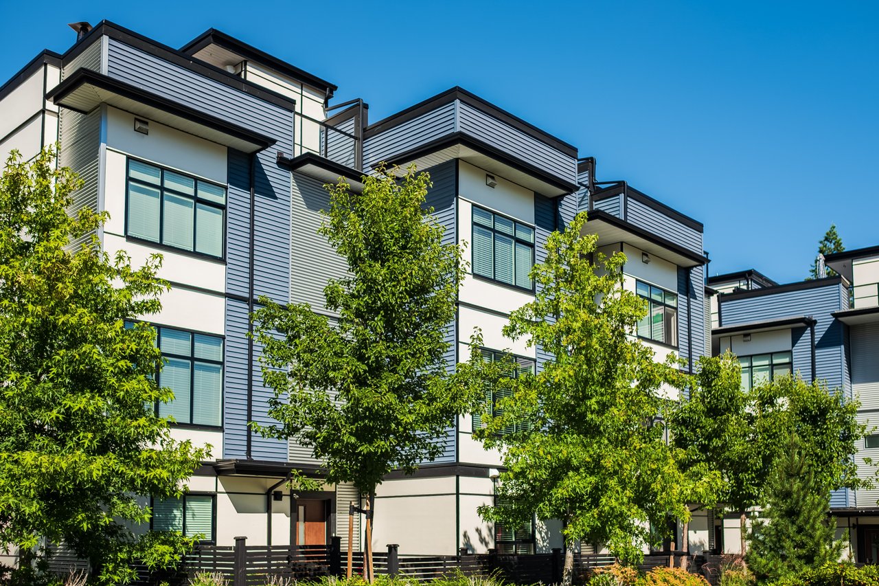 Modern condominium building with gray siding, dark trim, large windows, and leafy trees in front.