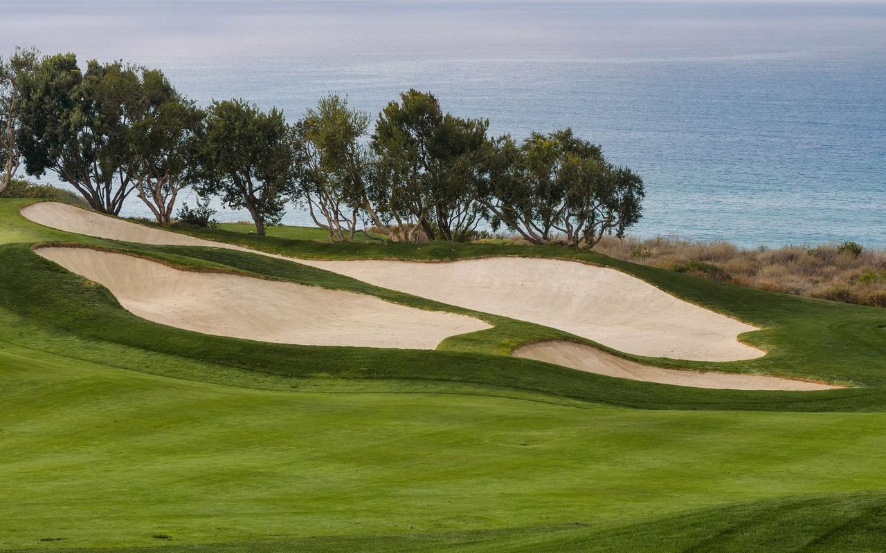 Coastal golf course with large sand bunkers, trees, and ocean beyond the fairway.