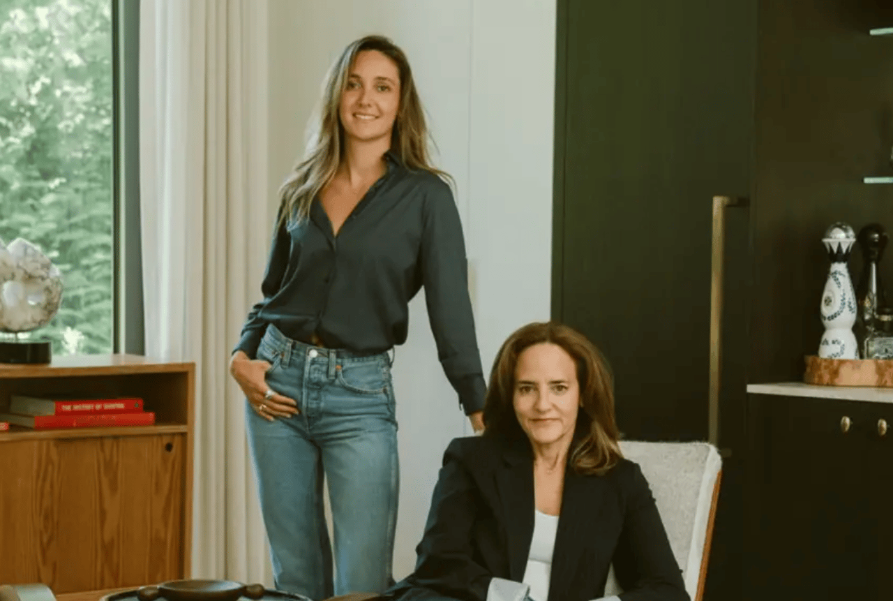 Two women pose in a modern interior, one standing and one seated.
