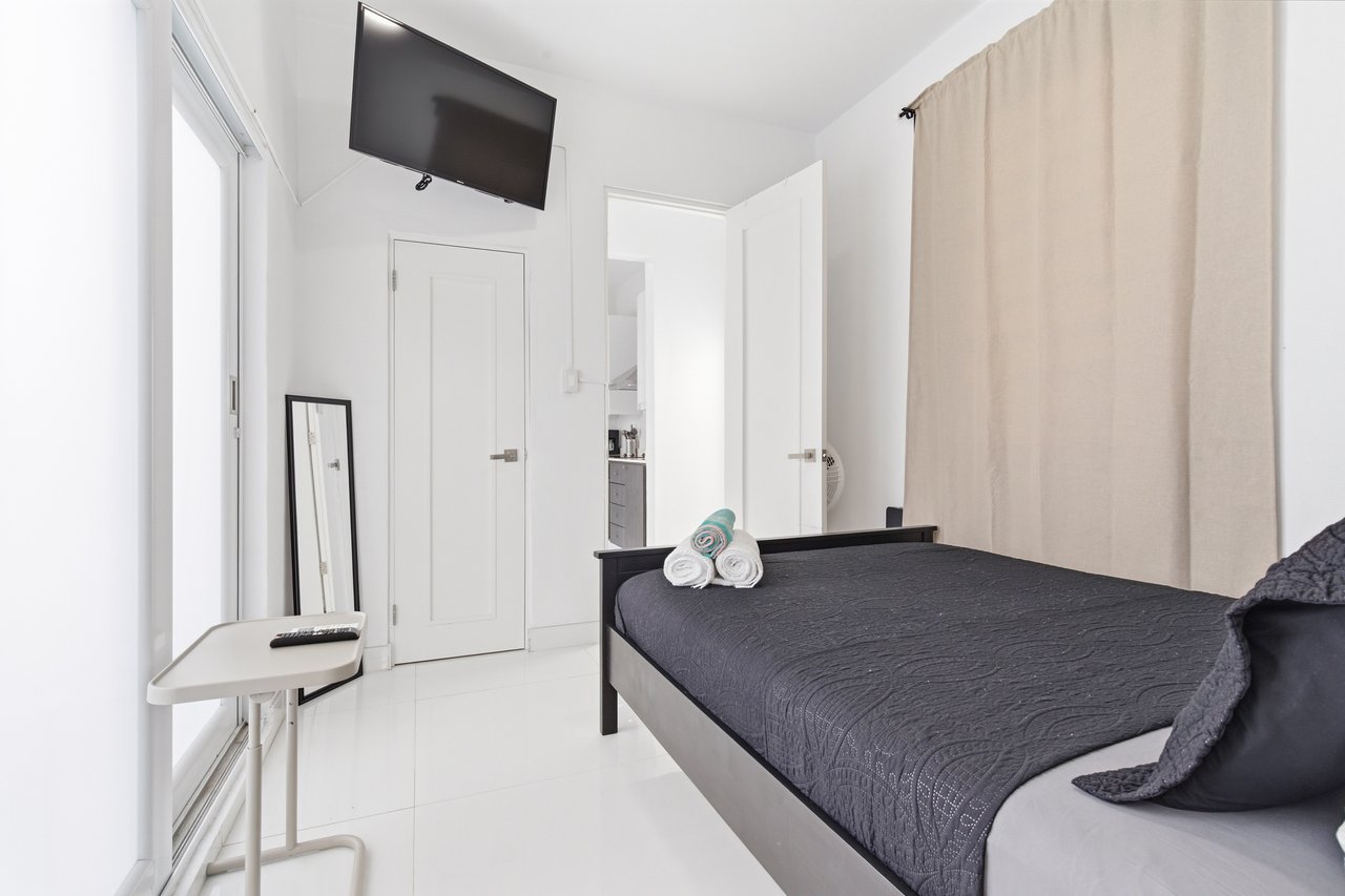 Furnished bedroom with a dark quilted bed, beige curtains, and a wall-mounted television.