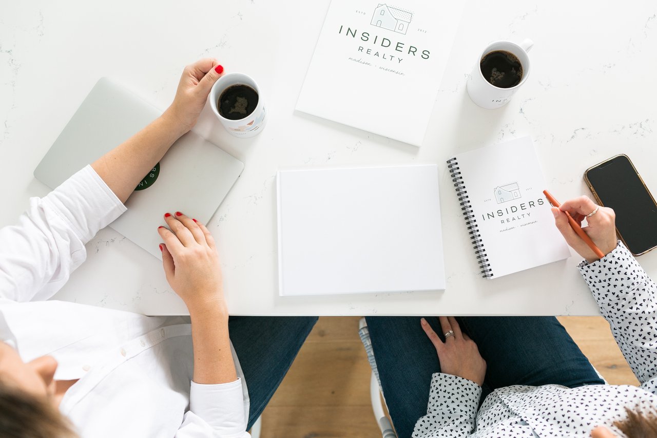 Overhead view of two people sitting at a white table with coffee, a laptop, and Insiders Realty branded notebooks, representing a strategy meeting to plan a successful home purchase with a low down payment in Sun Prairie, WI.