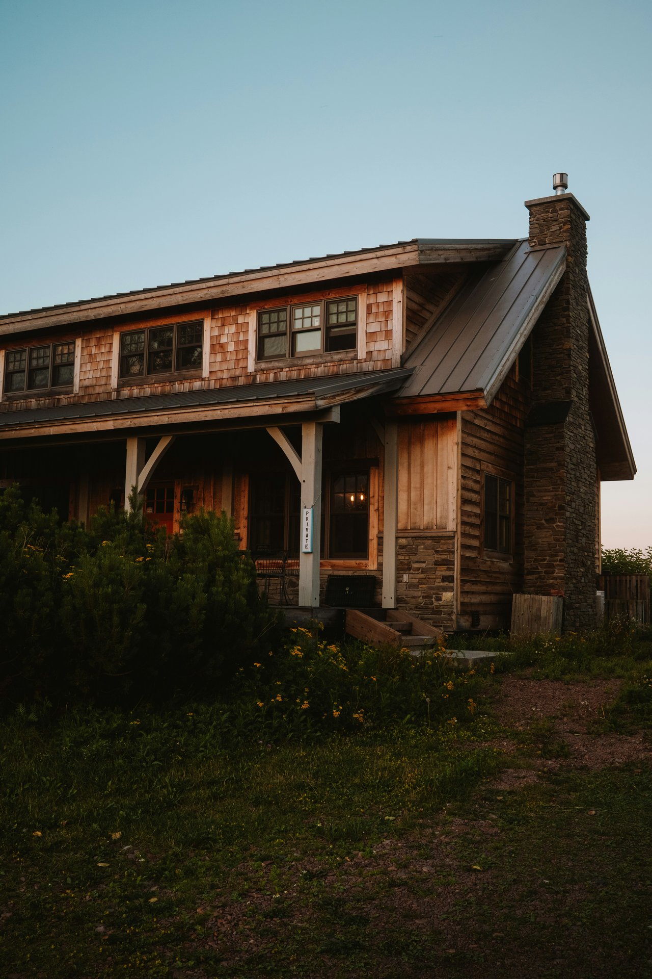 Rustic cedar-sided house with a stone chimney, metal roof, and covered porch at golden hour.