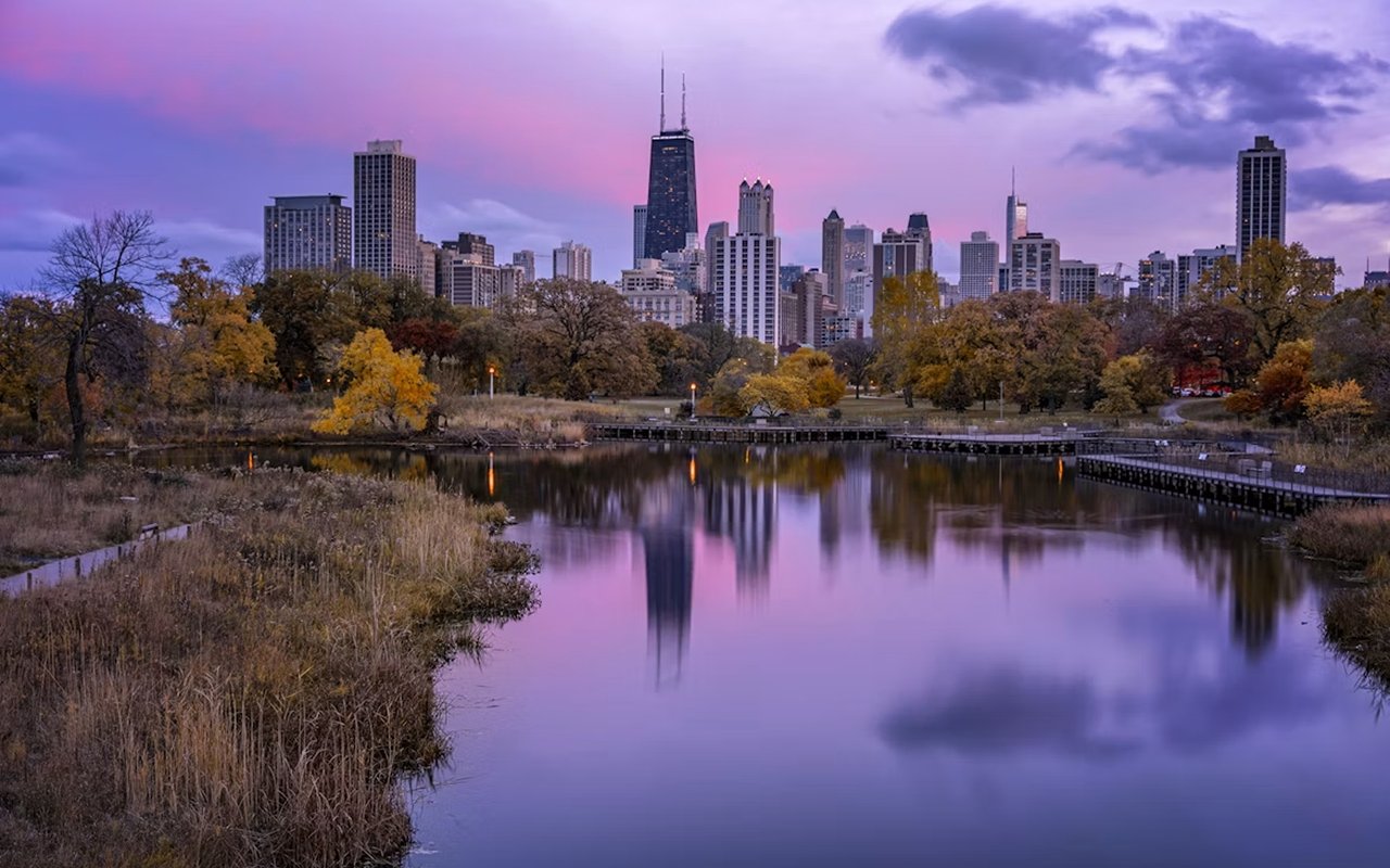 Chicago skyline reflected in a pond, framed by autumn trees beneath a pink and purple dusk sky.