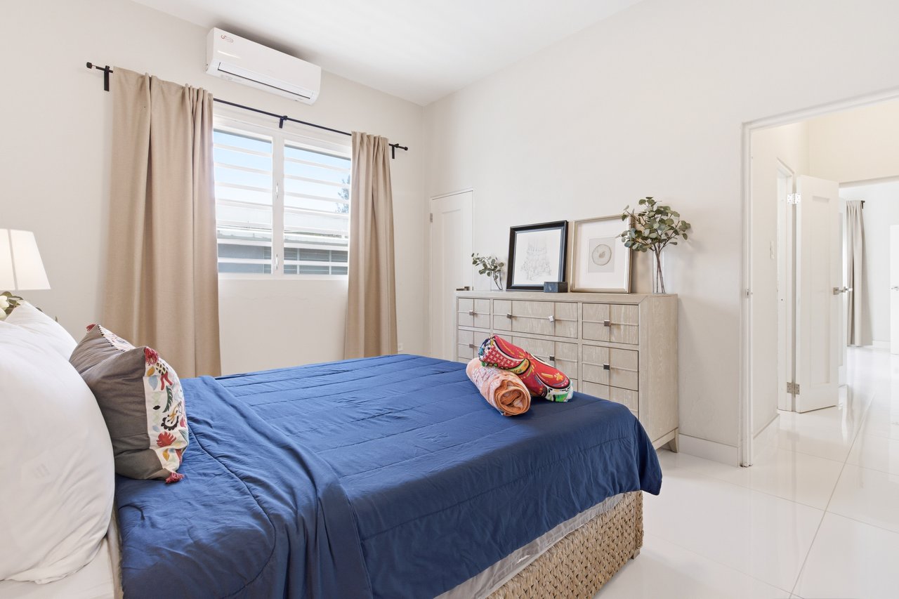 Bedroom with a blue-covered bed, beige curtains, and a dresser beneath framed art.