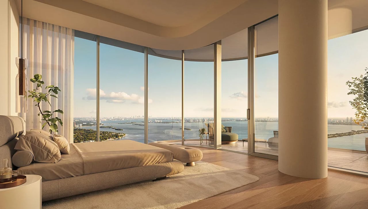 Staged bedroom with curved floor-to-ceiling windows overlooking water and a distant city skyline.