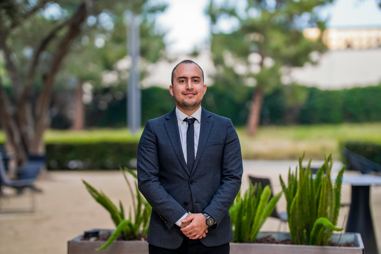 Sebastian Gomajoa in a navy suit and tie, standing outdoors among planters.