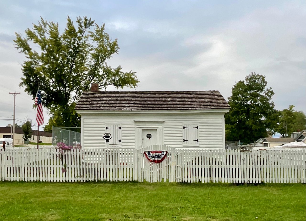 Small white clapboard building behind a picket fence, with an American flag and bunting.