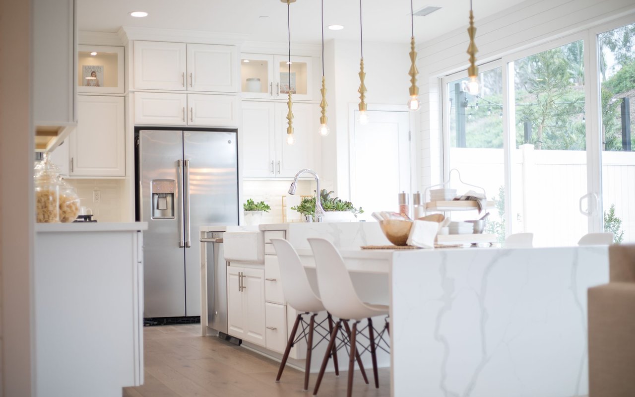 Bright white kitchen with a marble-look island, stainless steel refrigerator, and large windows.