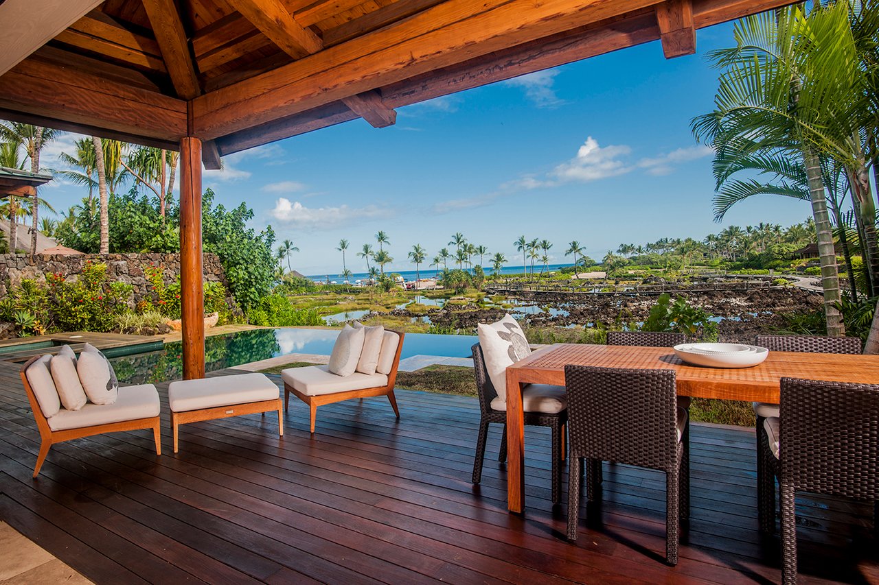 Covered lanai with outdoor dining and lounge furniture overlooking a pool, palms, and ocean.