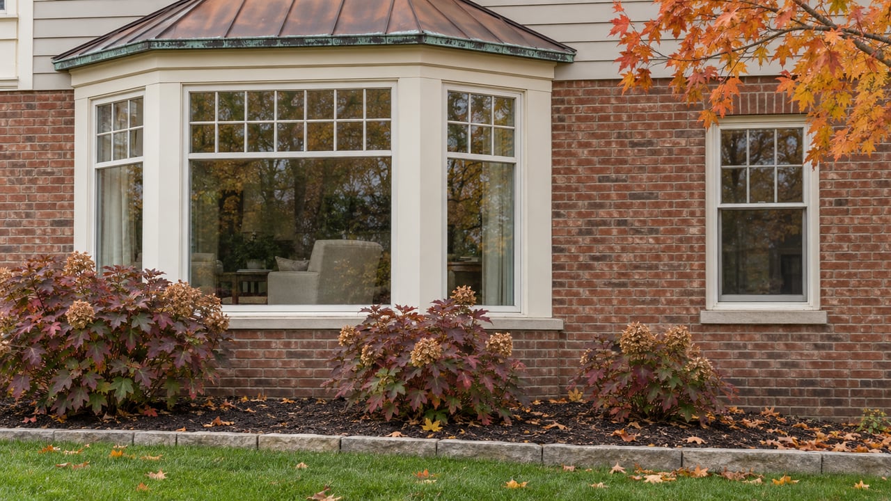 Copper-roofed ivory bay window on a brick home above oakleaf hydrangeas with autumn foliage.