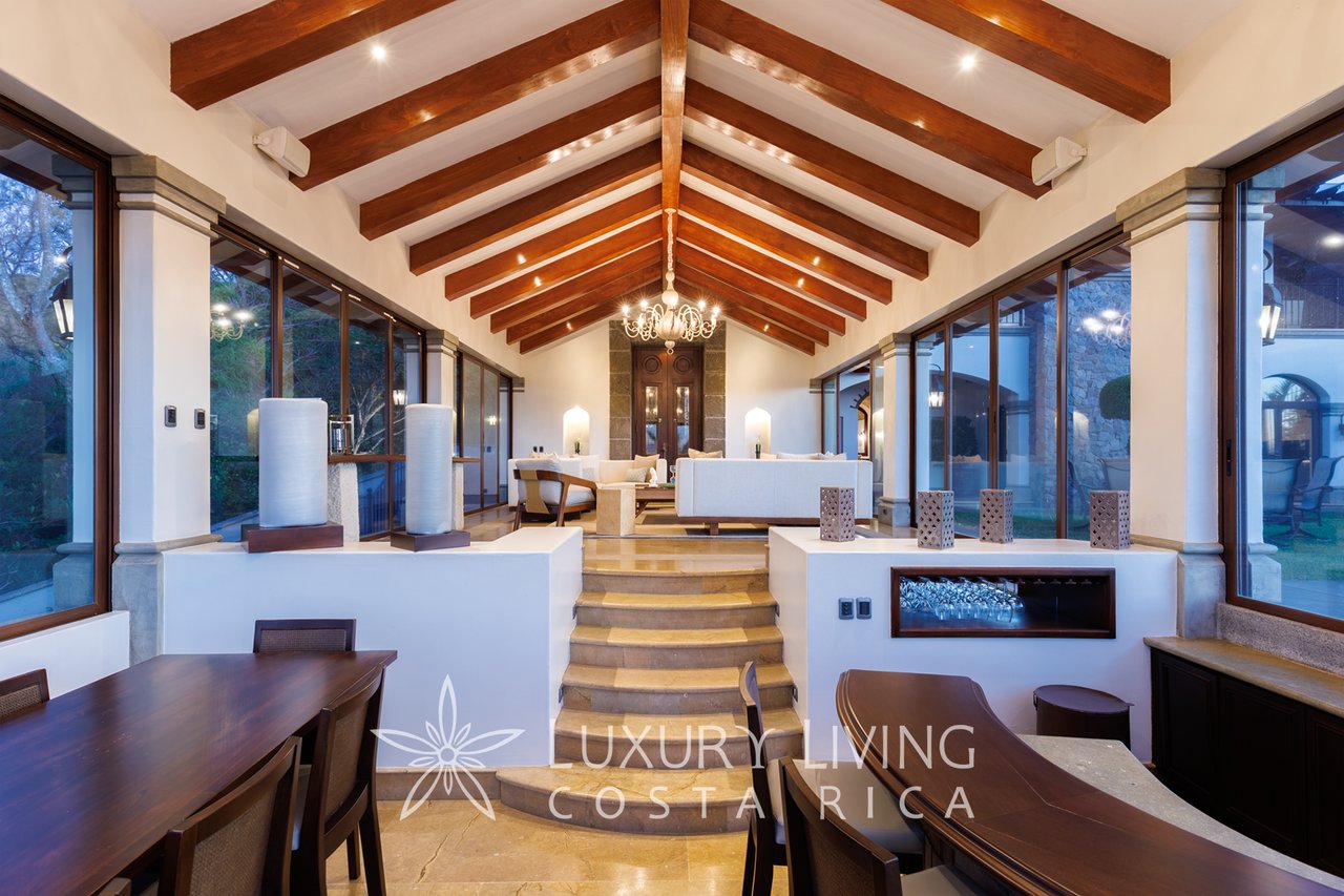 Furnished living and dining room with exposed timber beams, central steps, and glass walls.