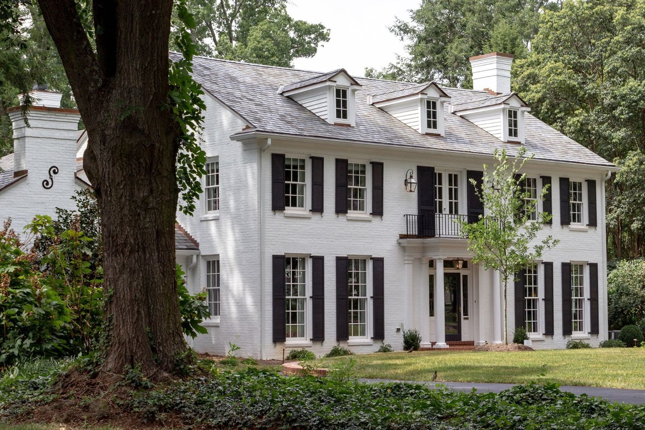 White brick two-story home with black shutters and a columned entry, framed by mature trees.