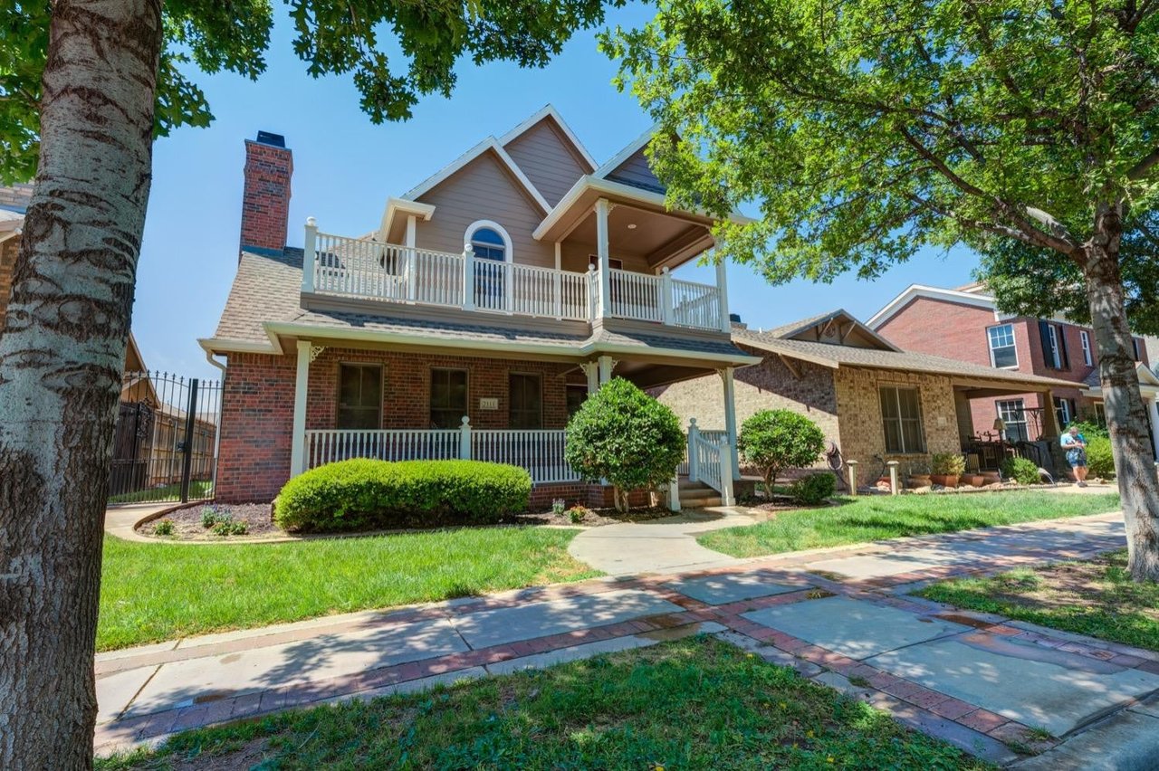 Two-story brick house with a white-railed balcony, covered porch, and leafy shade trees.