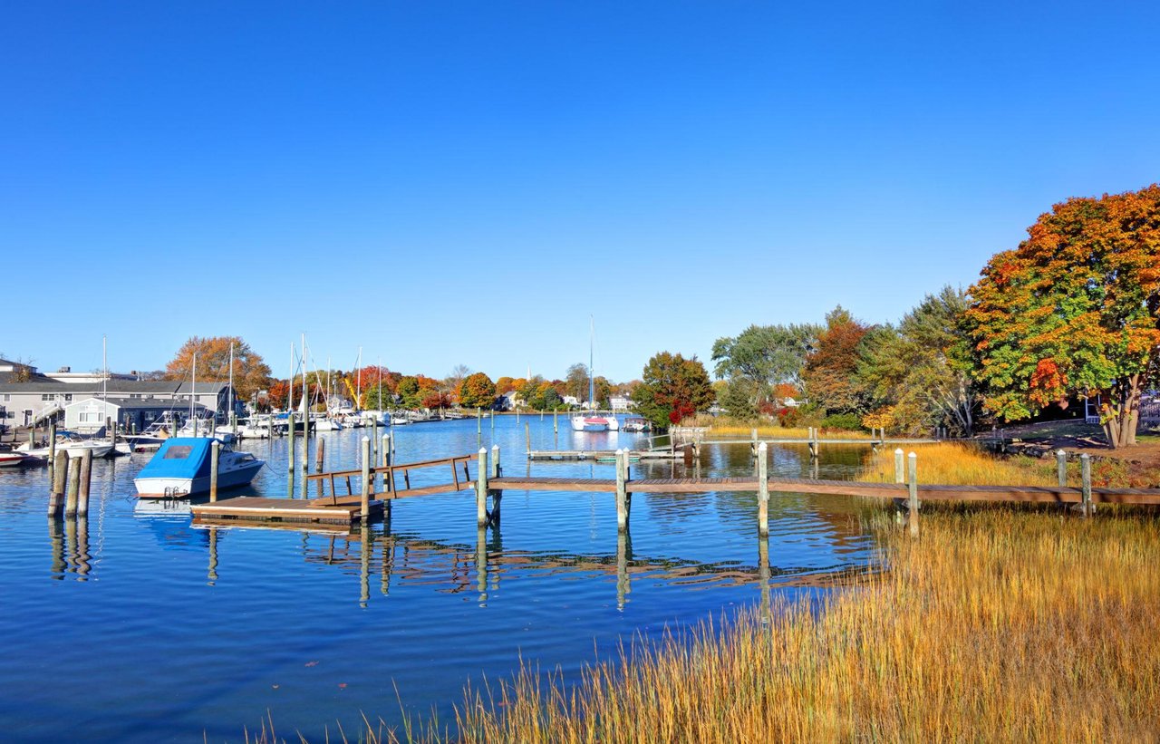Autumn harbor with boats and docks, blue water, marsh grasses, and colorful shoreline trees.