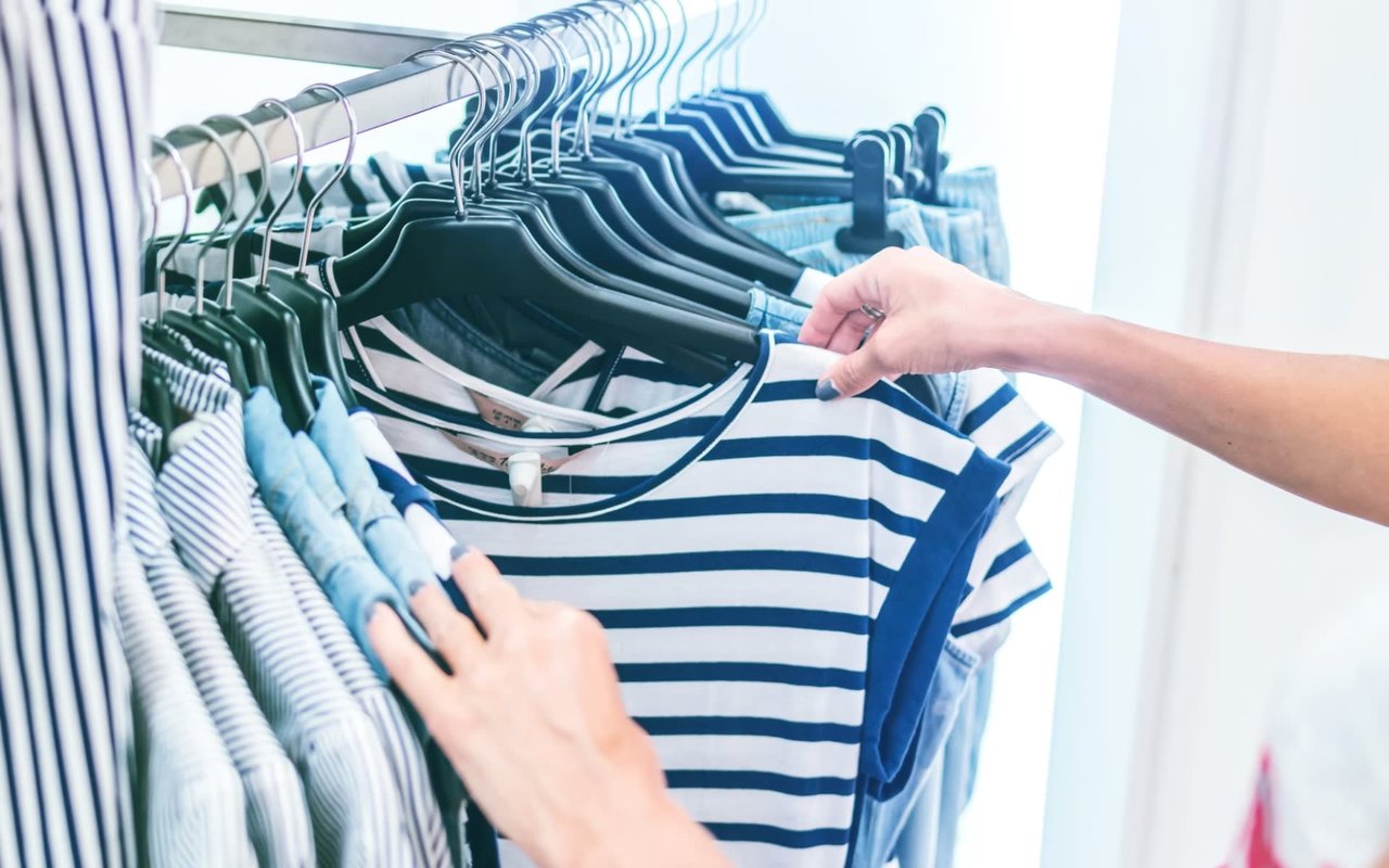 Close-up of a customer browsing a curated summer apparel collection, selecting a nautical blue and white striped t-shirt from a modern retail clothing rack.