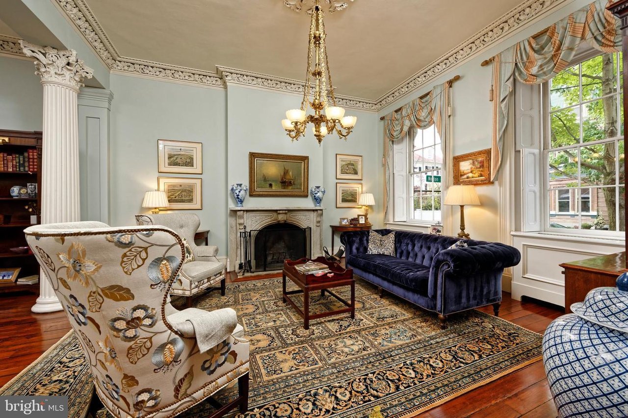 Formal living room with ornate ceiling molding, fireplace, chandelier, and tall street-facing windows.