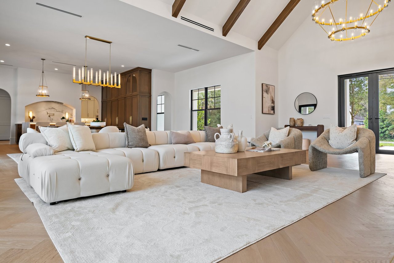 Open-plan living room with a white sectional, wood coffee table, vaulted beam ceiling, and black-framed glass doors.